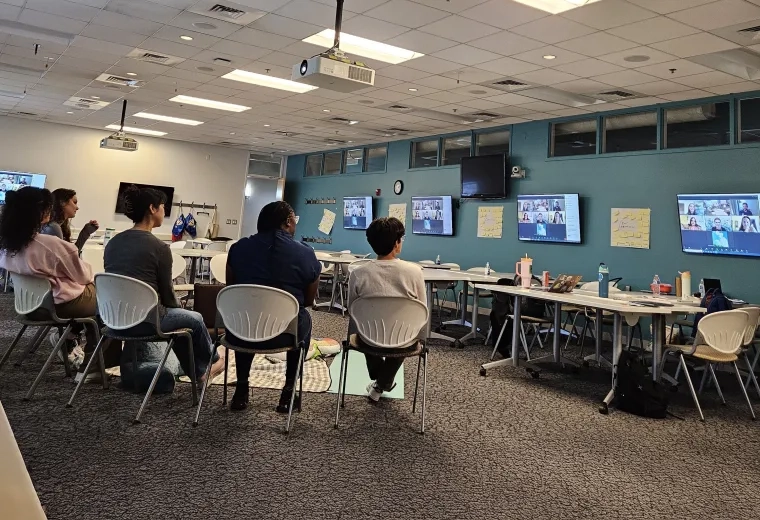 Six people sit in a semi circle watching screens with a virtual meeting