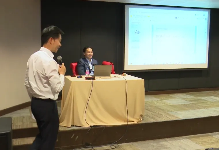 A man with a microphone stands in front of a table and screen