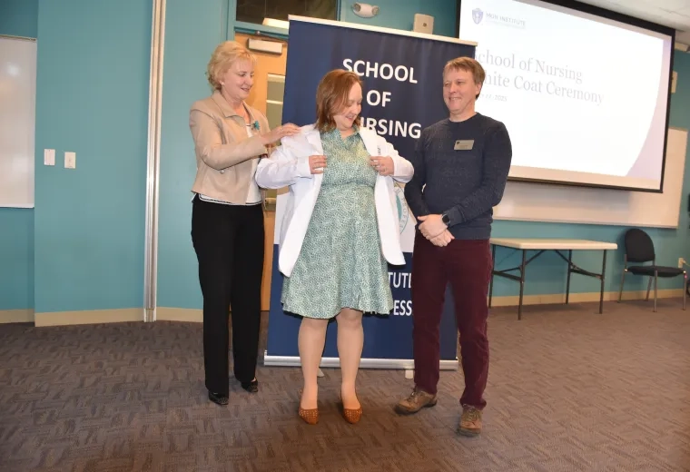 A woman helps another woman put on a white medical coat while a man observes