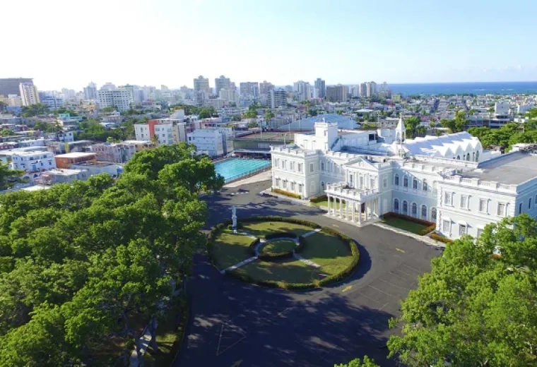 lush green trees frame a photo of a large white building with a city and the ocean in the background