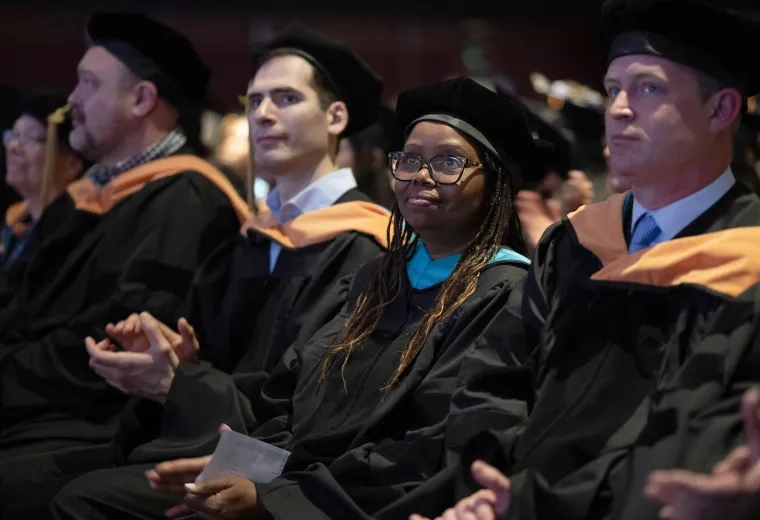 A woman wearing graduation robes sits in a crowd of people 