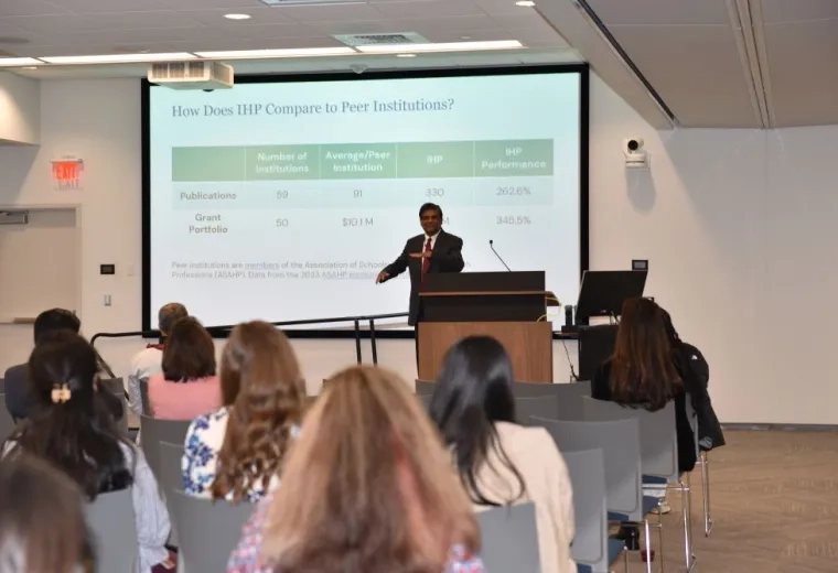 A man stands at a podium with a screen behind him that says "How does IHP Compare to Peer Institutions