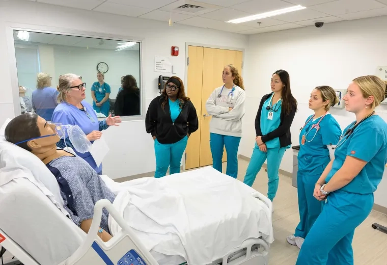a woman in scrubs gestures to a manikin in a hospital bed while other people in scrubs look on