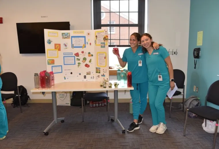 Two women wearing medical scrubs pose in front of a display board and table