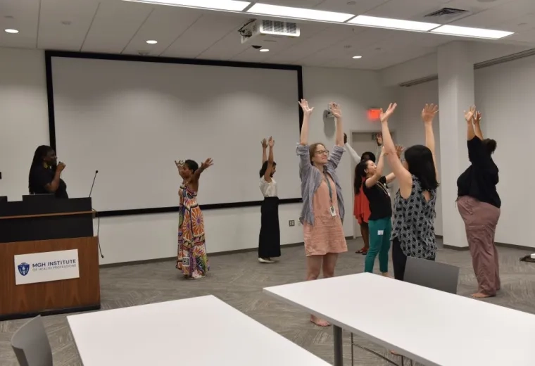 Several people stand facing different directions with their hands in the air while a woman with a microphone stands on the left