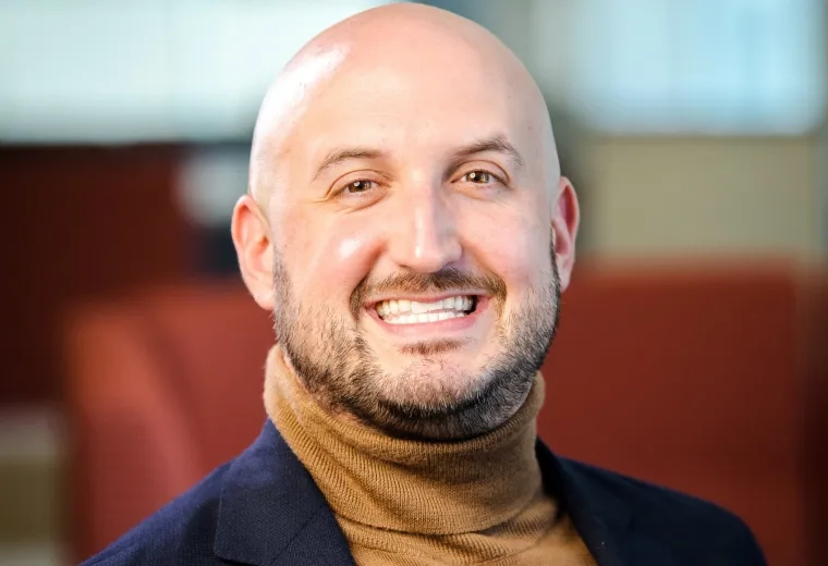 Greg Landry is bald and is wearing a brown turtleneck and navy blazer