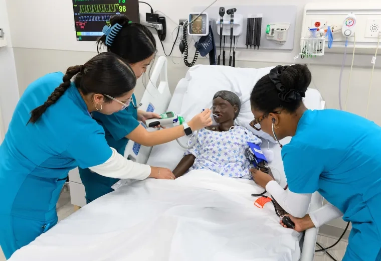 three women in blue scrubs surround a manikin child in a hospital bed