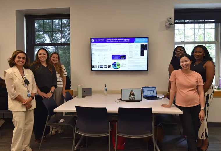 Six people stand around a table with a tv screen behind them showing a presentation