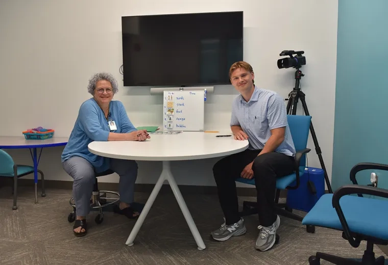 Two people sit at a table with a tv screen behind them 