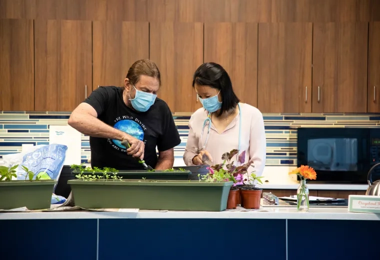 woman wearing mask and stethoscope helps middle aged man plant flowers in a pot