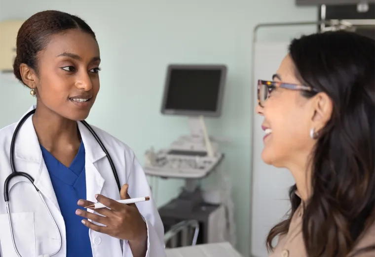 woman in a white coat and stethoscope speaks to another woman in an exam room