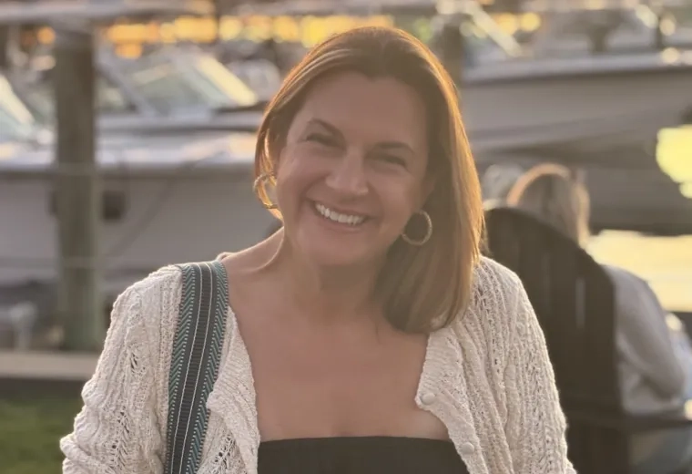 Woman wearing a black shirt and white sweater smiling and looking at camera. There is a sunset and boats on the water in the background.