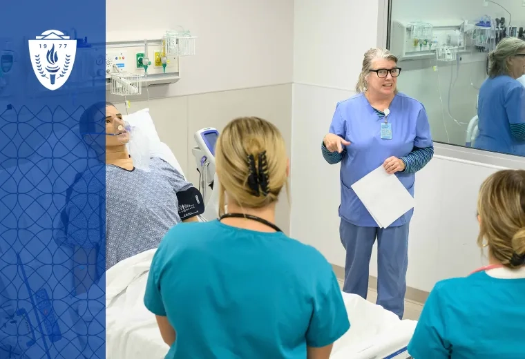 Professor and students wearing scrubs standing around a simulation hospital bed with a mannequin laying in the bed