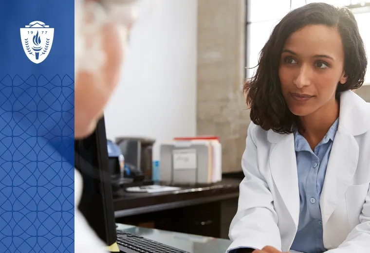 Woman wearing blue shirt and white lab coat sitting at desk accross from another person