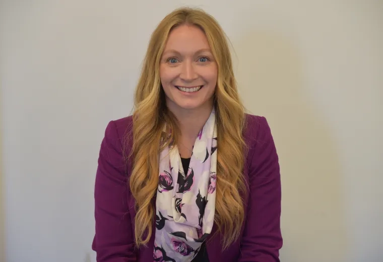 Headshot of woman with long blonde hair standing in front of solid white background wearing a light purple scarf and dark purple jacket