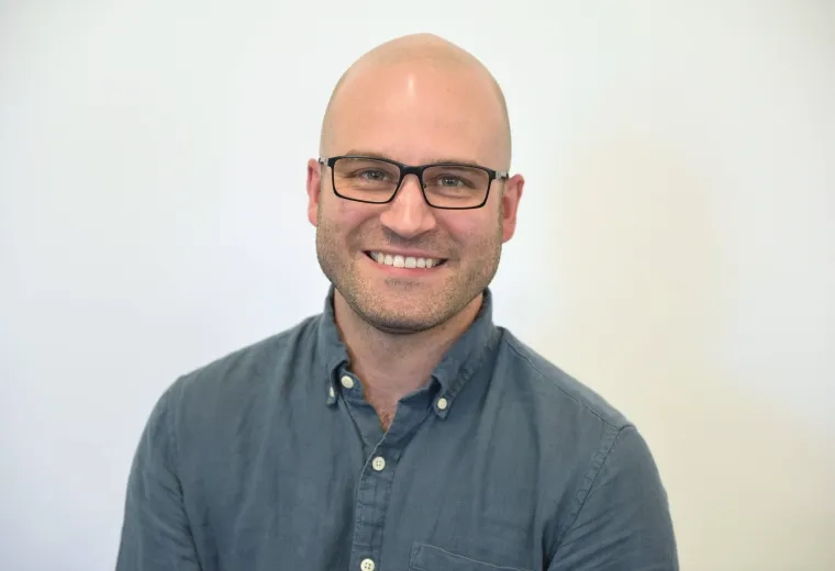 Headshot of man against a solid white background blue button-down shirt and glasses.