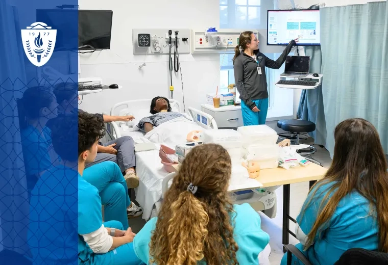 Group of students seated around simulation hospital bed with a mannequin laying on it. An instructor points toward the computer screen.