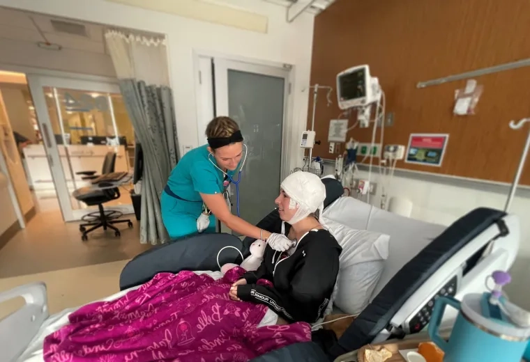 woman in scrubs holds a stethoscope to a woman's chest in a hospital bed