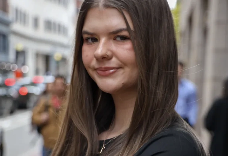 Headshot of woman with long brown hair wearing a black shirt in front of a city street in the background