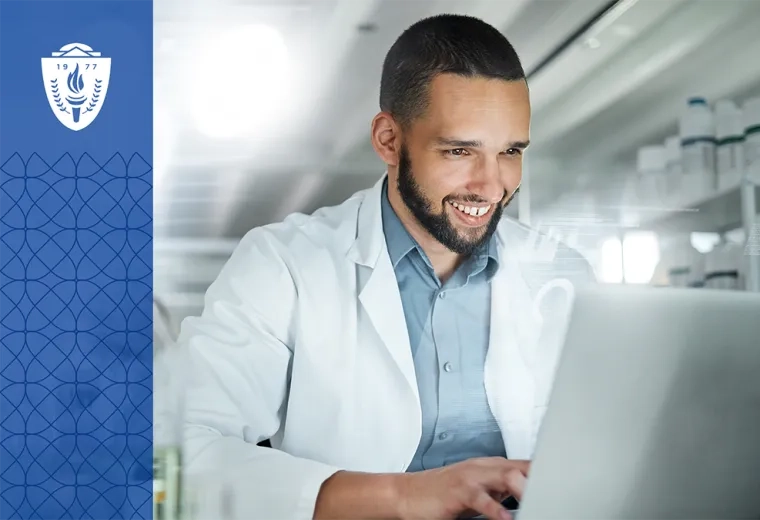 Man wearing blue shirt and white lab coat sitting in a lab and typing on a laptop.