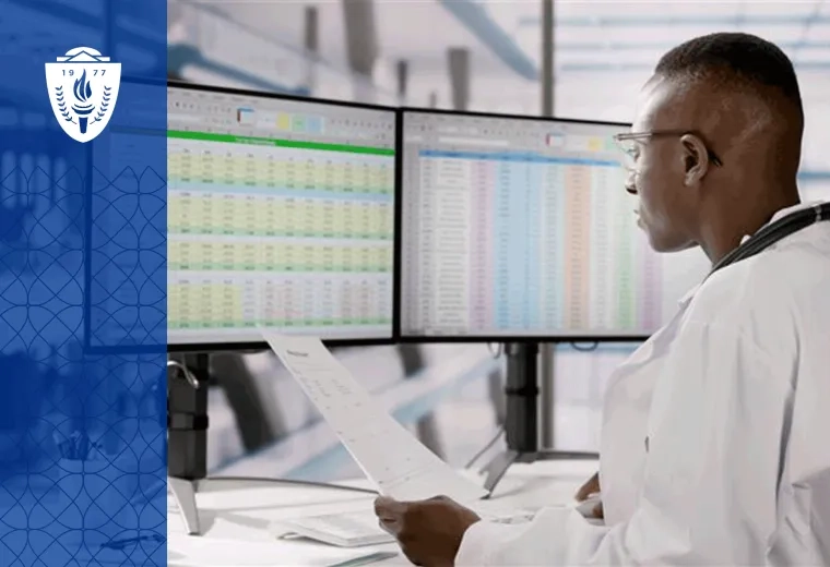 Man wearing lab coat sitting a desk and looking at spreadsheets on two computer monitors