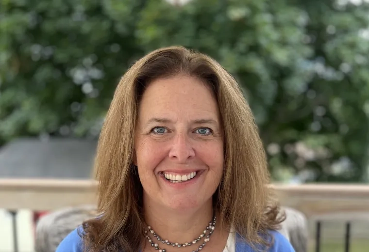 Headshot of woman with long brown hair wearing a white shirt, a blue sweater, and a necklace sitting on a balcony overlooking a lawn.