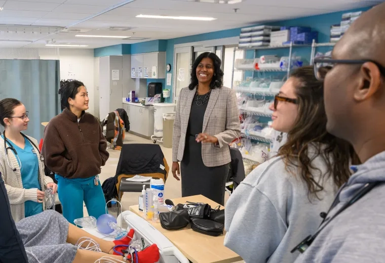 a woman speaks to nursing students in scrubs gathered around a hospital bed
