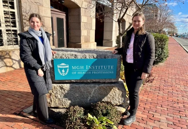Two women pose on either side of a rock with sign for MGH Institute