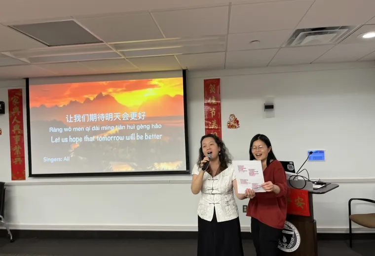 Two women sing in front of a screen with Chinese and English characters on it
