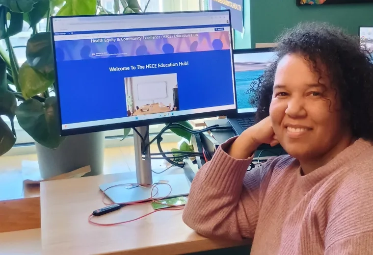 A woman looks at the camera with a computer screen behind her