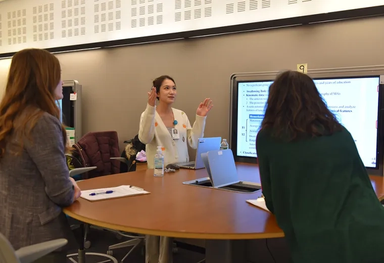 A woman stands in front of screen and talks to two people facing her