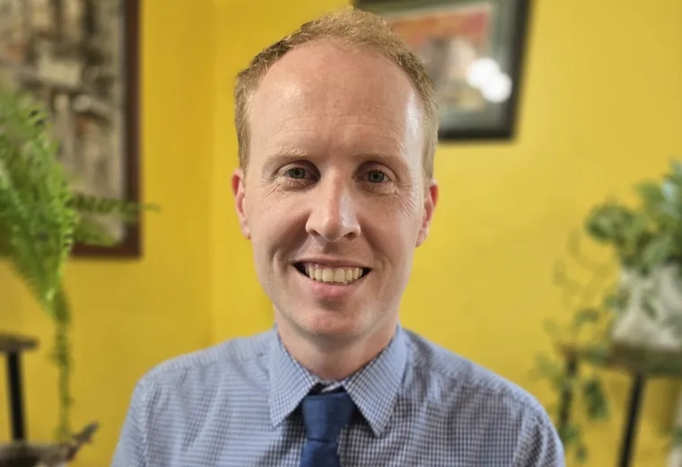 Headshot of Christopher Bodden wearing a blue button up shirt and blue tye in front of a yellow wall with paintings hung in the background.