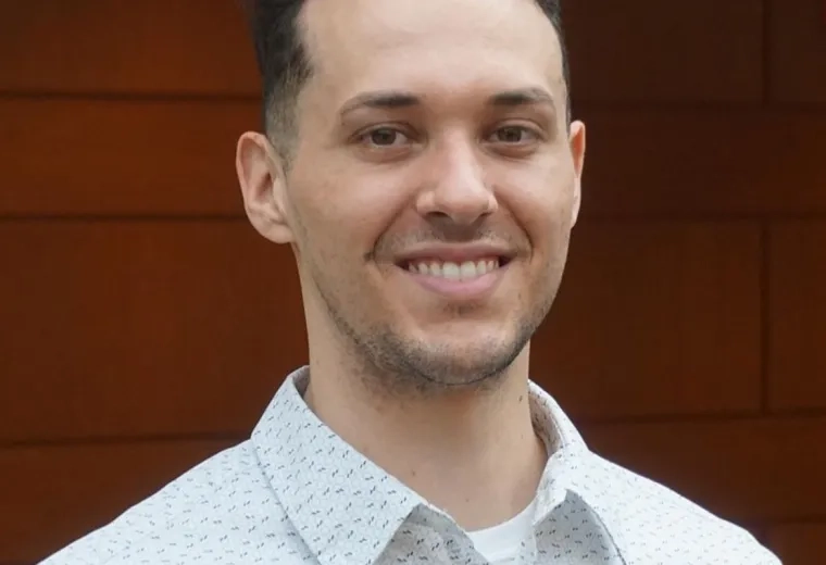 Man with short dark hair wearing a white patterned collared shirt in front of a red background