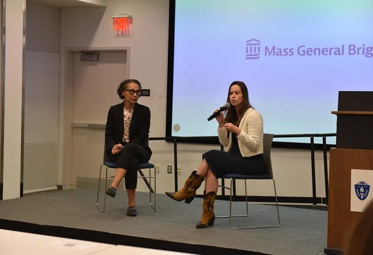 Two women sit on a stage with one speaking into a microphone