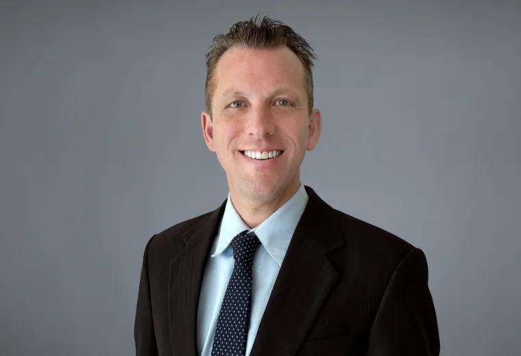 Headshot of Jason Richter standing against a solid gray background, wearing a blue collared shirt, a black blazer and, and blue necktie with white dots