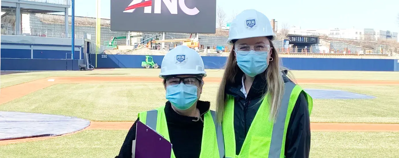 two women stand with ball field behind them wearing hard hats