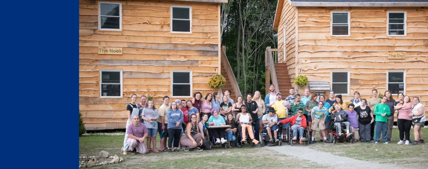 a large group of people stand and sit in wheelchairs in front of two wooden cabins