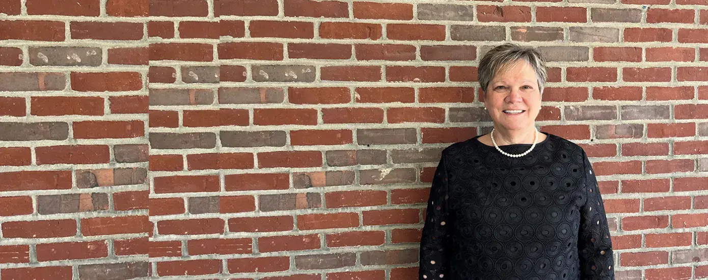 Doctor of Occupational Therapy program faculty member standing against a brick wall, smiling, and wearing a black blouse with a pearl necklace.
