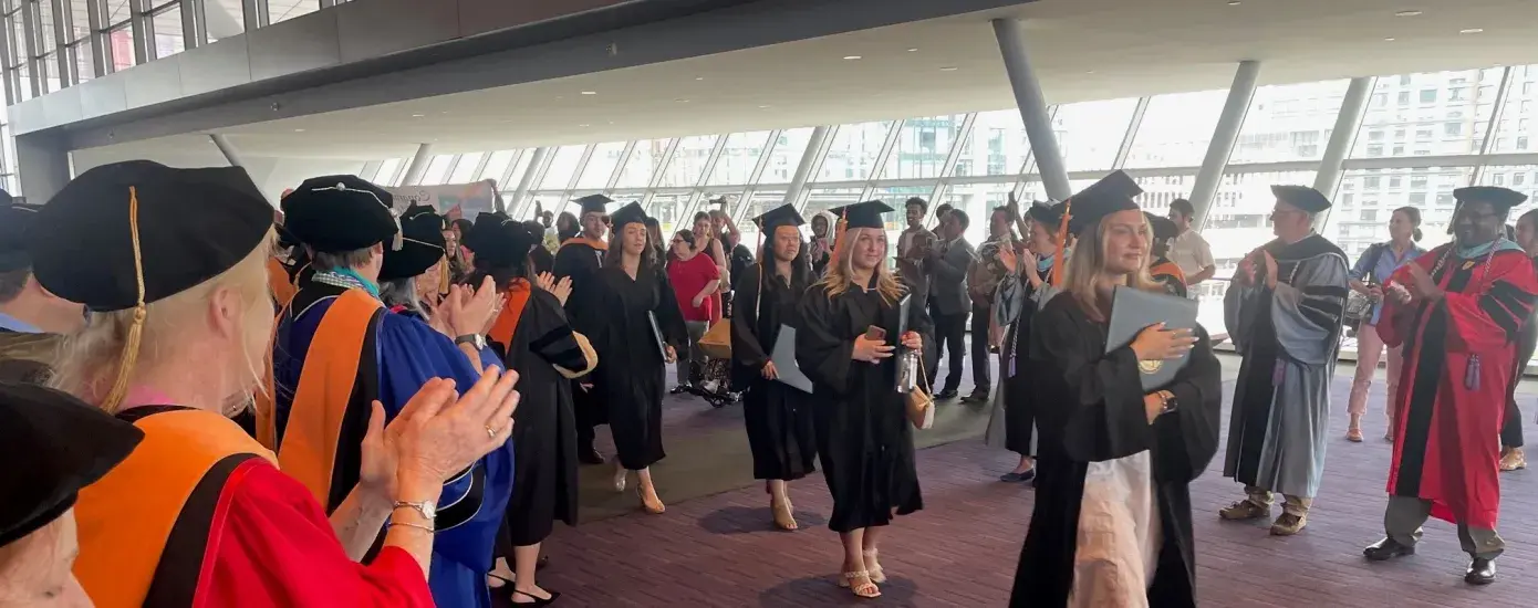 people in colorful graduation regalia clap as people wearing black caps and gowns walk in a line