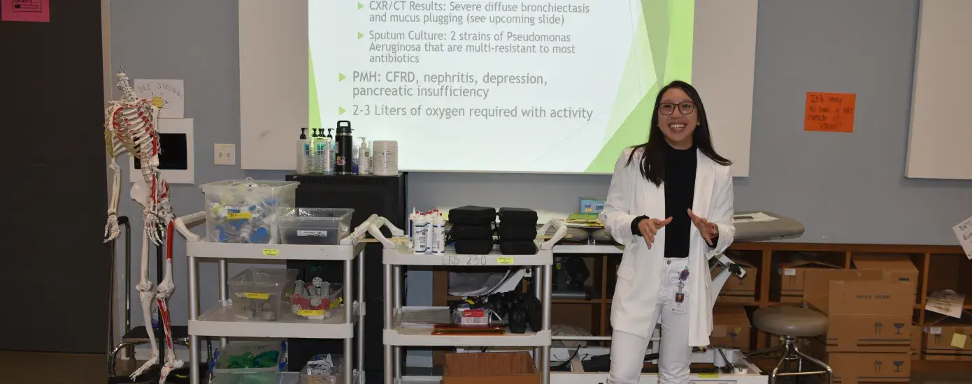 A woman in a white sweater and pants stands in front of a screen in a classroom with medical items behind her
