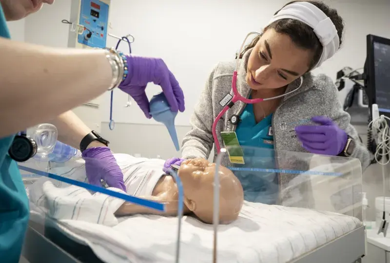 two women in scrubs look at a baby doll and use a suction bulb on it