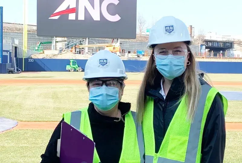 two women stand with ball field behind them wearing hard hats