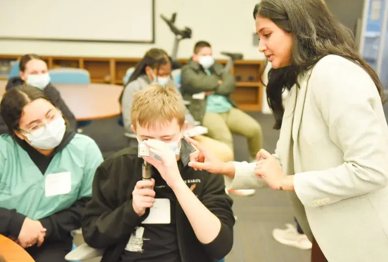 woman in blazer points at a cylindrical device that a man is holding to his eye