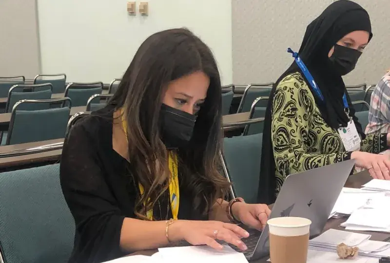 woman sits in chair at laptop next to another woman wearing a hijab
