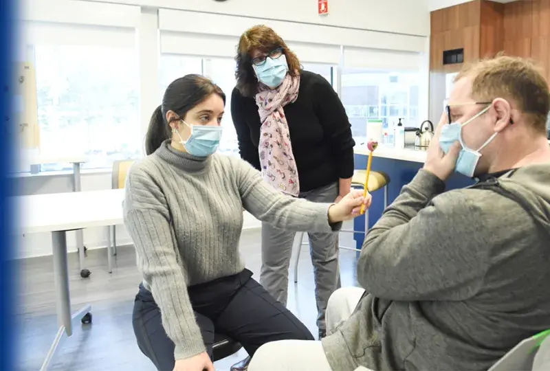 woman looks over the shoulder of a seated woman who holds a stick in front of the face of a man