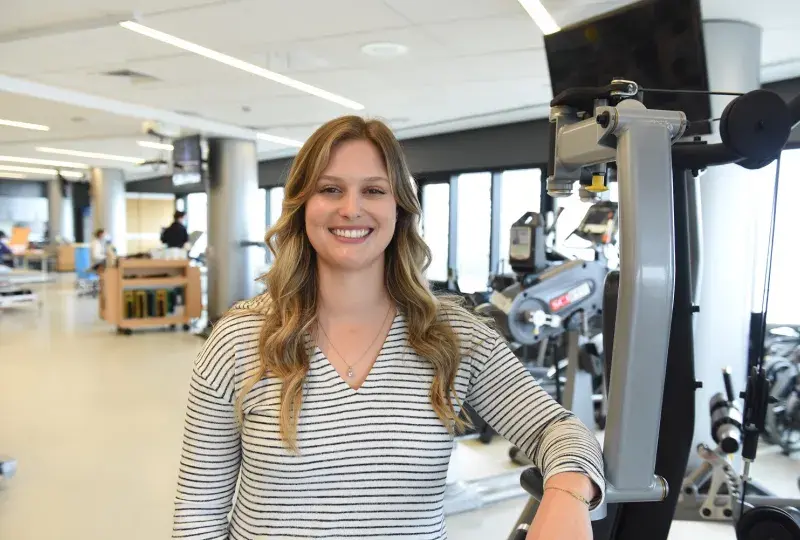Tarryn has wavy long dark blonde hair and wears a black and white striped top as she leans against a weight machine in a gym
