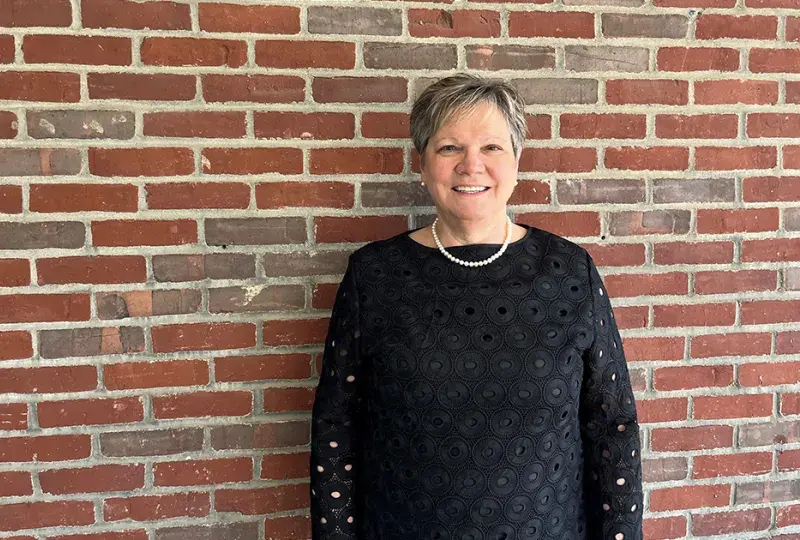 Doctor of Occupational Therapy program faculty member standing against a brick wall, smiling, and wearing a black blouse with a pearl necklace.