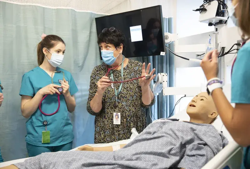 Karen holds a stethoscope and shows a student in scrubs how to apply it to a manikin in a hospital bed