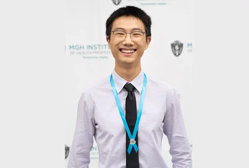 Asian man with black hair and glasses wears a black tie and off white collared shirt with a teal ribbon around his neck
