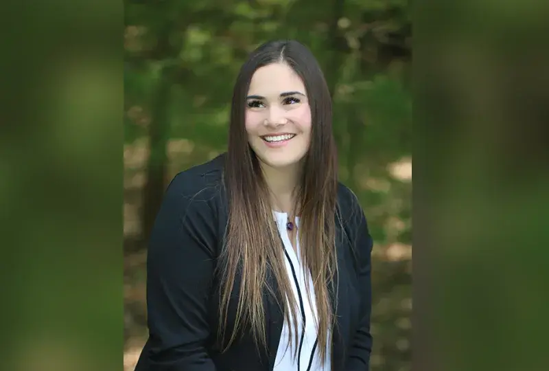 leah is outside in front of trees and she wears a white blouse under a black long sleeve top